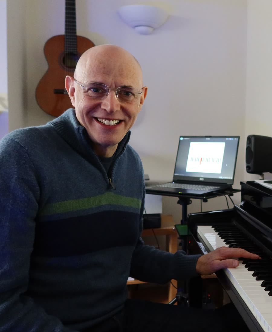 Robert Howard smiling at the piano in his music room, a guitar hanging on the wall behind him